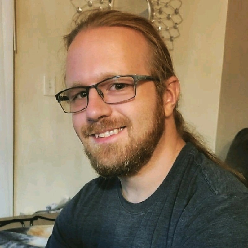 Jeremy Matthews smiling at the camera, with glasses, a full beard, and long hair tied back, wearing a dark gray t-shirt